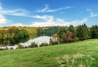 Vista desde la casa móvil Manureva en Bélgica con prado verde, lago y colinas arboladas bajo cielo azul.