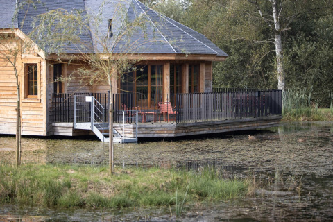 Wooden lodge named 'Happiness' in Belgium, featuring a deck over the water and surrounded by nature.