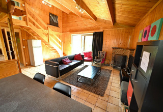 Salon en bois du lodge Ard Zen en Belgique avec canapé moderne, cheminée et escalier ouvert.