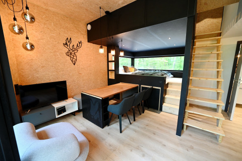 Modern lodge interior with elevated bed, wooden stairs, dining area, and forest view in Belgium.