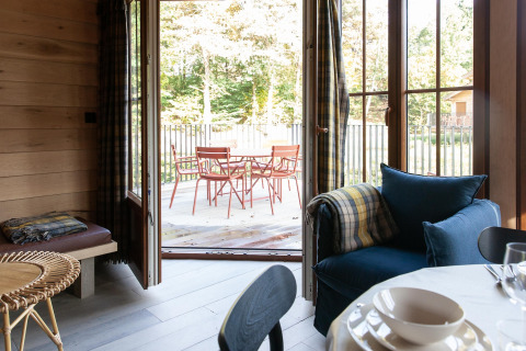 Interior view from Cabane 'The Funny Friends' lodge in Belgium with open doors leading to a terrace and garden chairs.