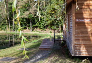 A wooden lodge named 'The Funny Friends' by a pond, surrounded by trees, Belgium, sunny weather.