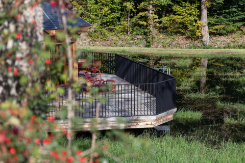 Udsigt over en træveranda med sorte rækværk og havemøbler ved en sø, omgivet af skov ved Cabane «The Funny Friends» i Belgien.