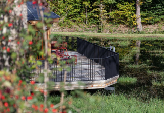 Terraza de madera con barandilla negra y sillas junto a un estanque, en medio del bosque en Cabane «The Funny Friends», Bélgica.