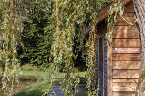 Træhytte ved navn 'The Funny Friends' ved Cabane i Belgien, omgivet af grønne træer og natur.
