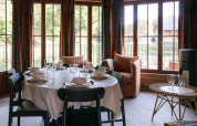 Sala da pranzo in un lodge belga con tavola apparecchiata, ampie finestre e vista su baite di legno fuori.