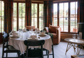 Sala da pranzo in un lodge belga con tavola apparecchiata, ampie finestre e vista su baite di legno fuori.