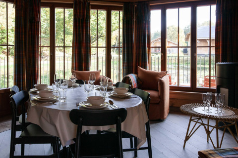 Dining room in a Belgian lodge with a set table, large windows, and a view of wooden cabins outside.