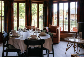 Dining room in a Belgian lodge with a set table, large windows, and a view of wooden cabins outside.