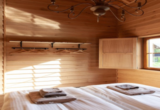 Bright wooden bedroom at lodge 'Happiness', Belgium, with bed, towels, coat rack and sunlight.