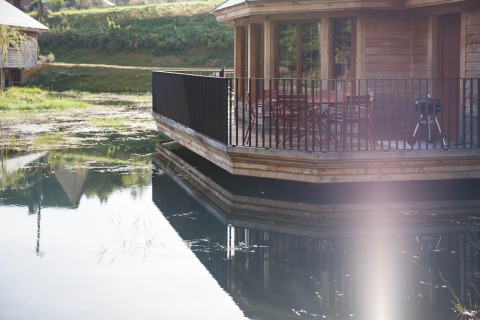 Wooden lakeside lodge named Happiness in Belgium, featuring a terrace with chairs and reflections on the water.