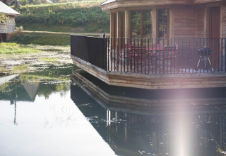Wooden lakeside lodge named Happiness in Belgium, featuring a terrace with chairs and reflections on the water.