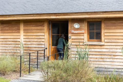 Person entering a wooden lodge named 'Happiness' with plants in front, photo taken in Belgium.