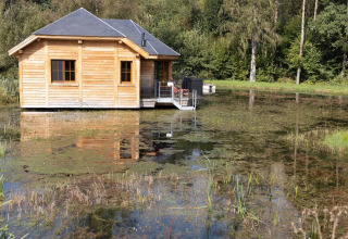 Wooden lodge named 'Happiness' on a pond in Belgium, surrounded by nature with reflection in the water.
