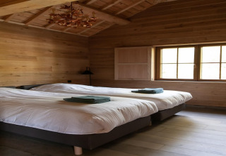 Room at lodge ‘Happiness’ in Belgium featuring two single beds, wooden walls, and sunlight through window.