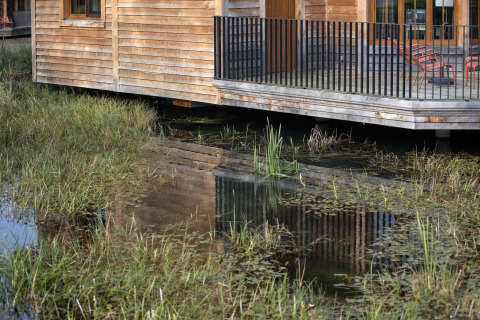 A wooden lakeside lodge called “Happiness” in Belgium, with porch and water reflection in the foreground.