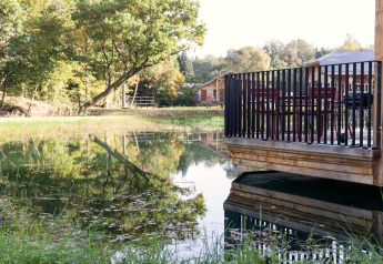 Terrazza in legno di un lodge sul lago in Belgio, con barbecue, sedie e alberi riflessi nell'acqua.