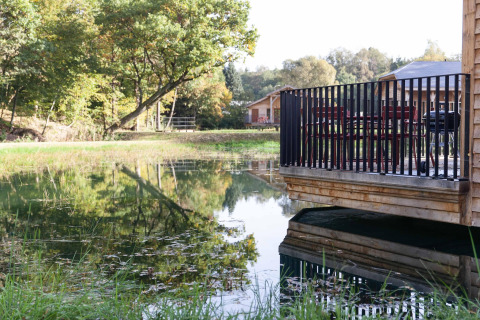 Wooden deck by a lakeside lodge in Belgium, featuring a grill, chairs and trees reflected in the calm water.