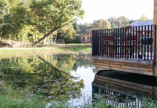 Wooden deck by a lakeside lodge in Belgium, featuring a grill, chairs and trees reflected in the calm water.