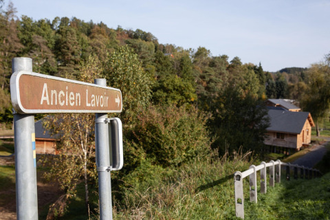A sign reading 'Ancien Lavoir' points toward a wooden lodge named 'Happiness' in Belgium's greenery.