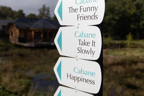 Signposts reading 'Cabane The Funny Friends', 'Take It Slowly' and 'Happiness' near a Belgian lodge over water.