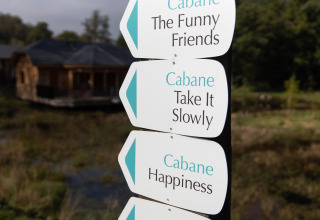 Signposts reading 'Cabane The Funny Friends', 'Take It Slowly' and 'Happiness' near a Belgian lodge over water.