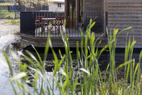 View of a wooden deck at lodge 'Happiness' in Belgium, featuring tables, chairs, water, and reeds.