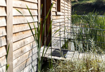 Ingresso alla lodge 'Happiness' in Belgio con pareti in legno, canneti e ambiente naturale acquatico.