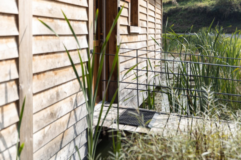 Entrance of the 'Happiness' lodge in Belgium with wooden siding, reeds, and natural pond scenery.