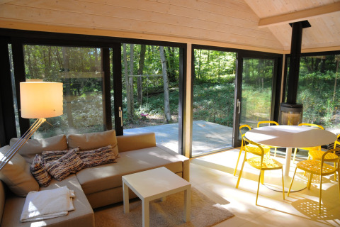 Licht chaletinterieur met grote ramen, beige bank, gele stoelen en uitzicht op het bos in België.