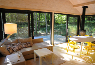 Licht chaletinterieur met grote ramen, beige bank, gele stoelen en uitzicht op het bos in België.