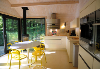 Cocina de chalet moderna con sillas amarillas, mesa redonda y grandes ventanas hacia el bosque en Bélgica