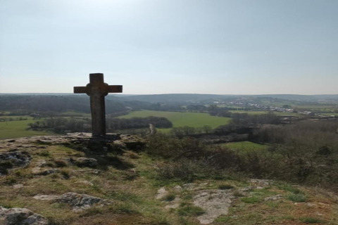Croce di pietra su una collina vicino a uno chalet con vista su campi verdi e un villaggio lontano.