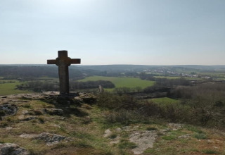 Croce di pietra su una collina vicino a uno chalet con vista su campi verdi e un villaggio lontano.