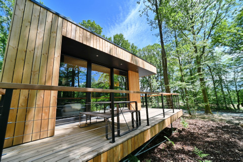 Modern wooden lodge with spacious deck set amidst green forest, sunny sky, Village de Vacances Oignies 5, Belgium.