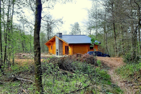 Chalet in legno al Village de Vacances Oignies 6 immerso tra gli alberi, con un’auto nera parcheggiata vicino.