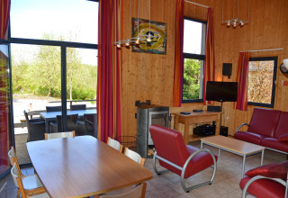 Lys stue i et træbeklædt sommerhus med røde møbler, store vinduer og udsigt til terrasse og natur.