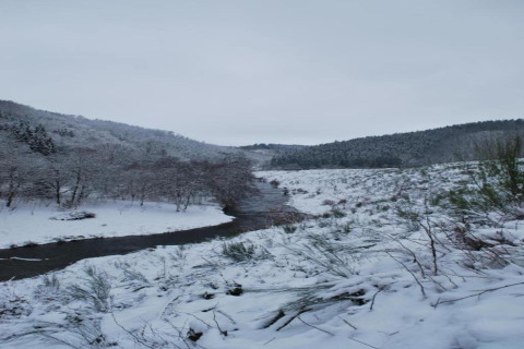 Snedækket landskab ved en flod nær Les Jardins de l'Ourthe 8 i Belgien, omgivet af bakker og skov.