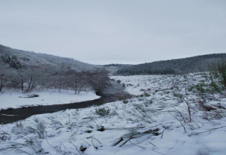 Snedækket landskab ved en flod nær Les Jardins de l'Ourthe 8 i Belgien, omgivet af bakker og skov.
