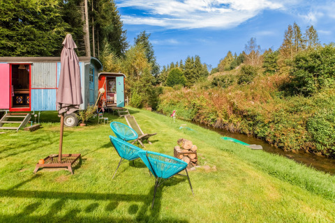 Les Roulottes de Vio mobile home in Belgium with colorful wagons, chairs, and a small stream on green grass.