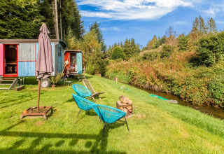 Les Roulottes de Vio mobile home in Belgium with colorful wagons, chairs, and a small stream on green grass.