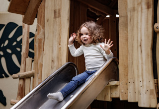 Kind met krullen glijdt vrolijk van een houten glijbaan bij een glampingaccommodatie.
