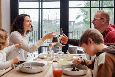 Gezin geniet samen van het diner en wijn in een moderne glamping accommodatie met grote ramen.