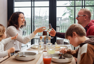 Gezin geniet samen van het diner en wijn in een moderne glamping accommodatie met grote ramen.