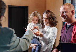Een gezin met een jong kind checkt in bij de receptie van een glampingaccommodatie en lacht vriendelijk.