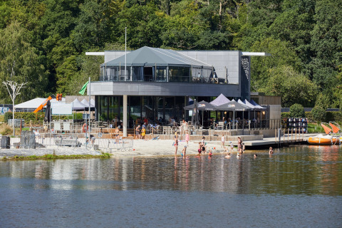 Glamping-accommodatie aan een meer, mensen zwemmen en ontspannen bij een modern restaurant.