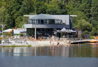 Glamping-accommodatie aan een meer, mensen zwemmen en ontspannen bij een modern restaurant.