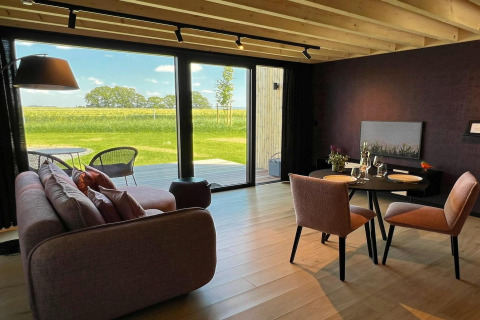 Lys stue i en hytte ved navn 'Natural Escape' i Belgien med udsigt til grønne marker gennem store vinduer.