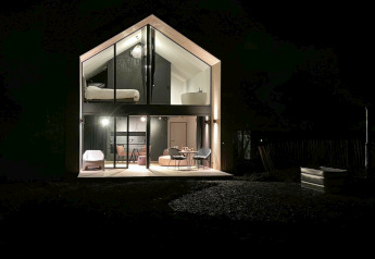 Foto nocturna de la cabaña 'Natural Escape' en Bélgica, fachada moderna de cristal y cálida iluminación.
