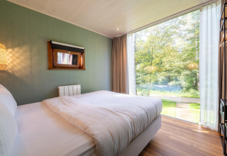 Schlafzimmer im TreeLoft von YourNature mit Panoramafenster und Blick auf einen grünen, bewaldeten Außenbereich.